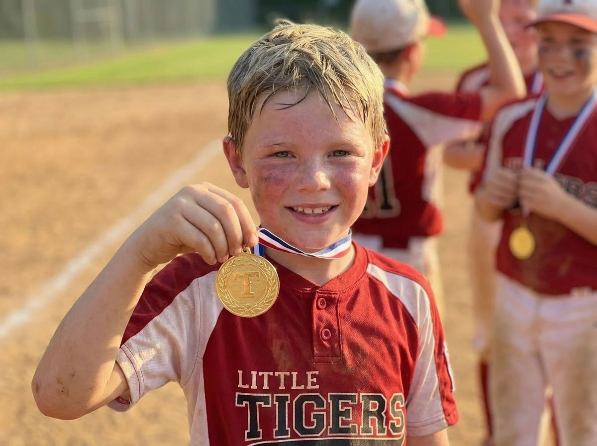 Young Little Tigers baseball player smiling and proudly holding up a gold medal on the baseball field after winning a tournament.