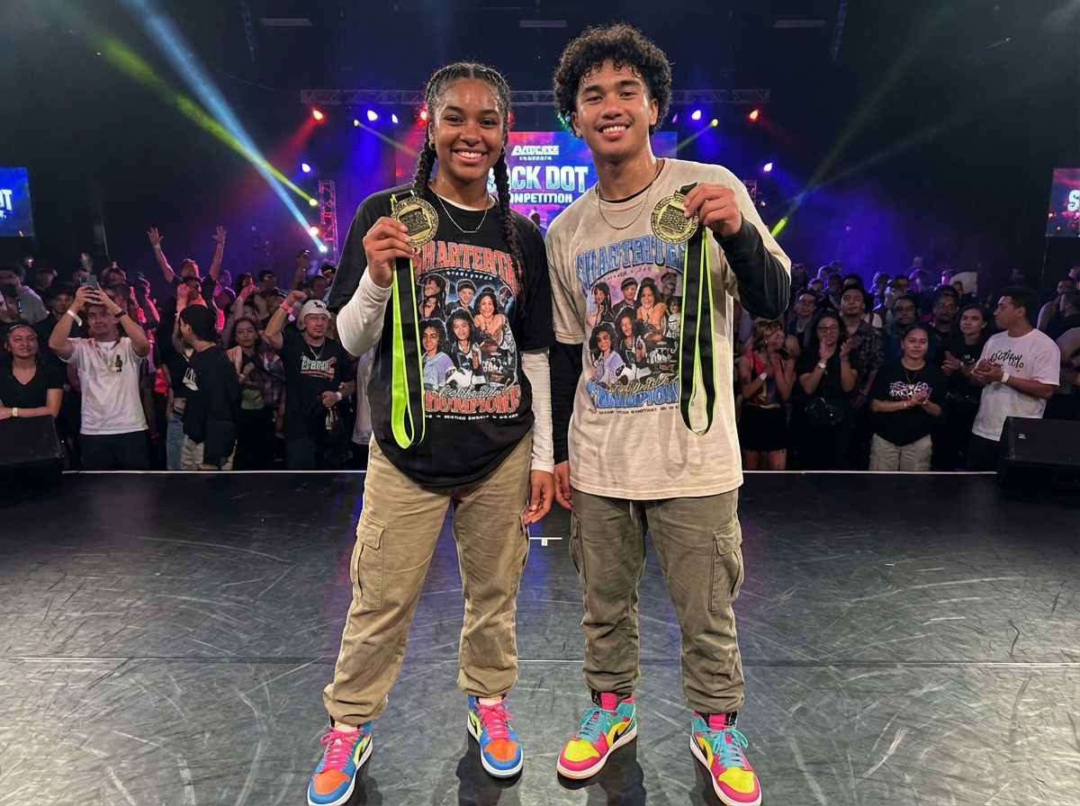 Two hip-hop dancers on stage holding up gold medals on neon green ribbons after winning a Black Dot street dance competition.
