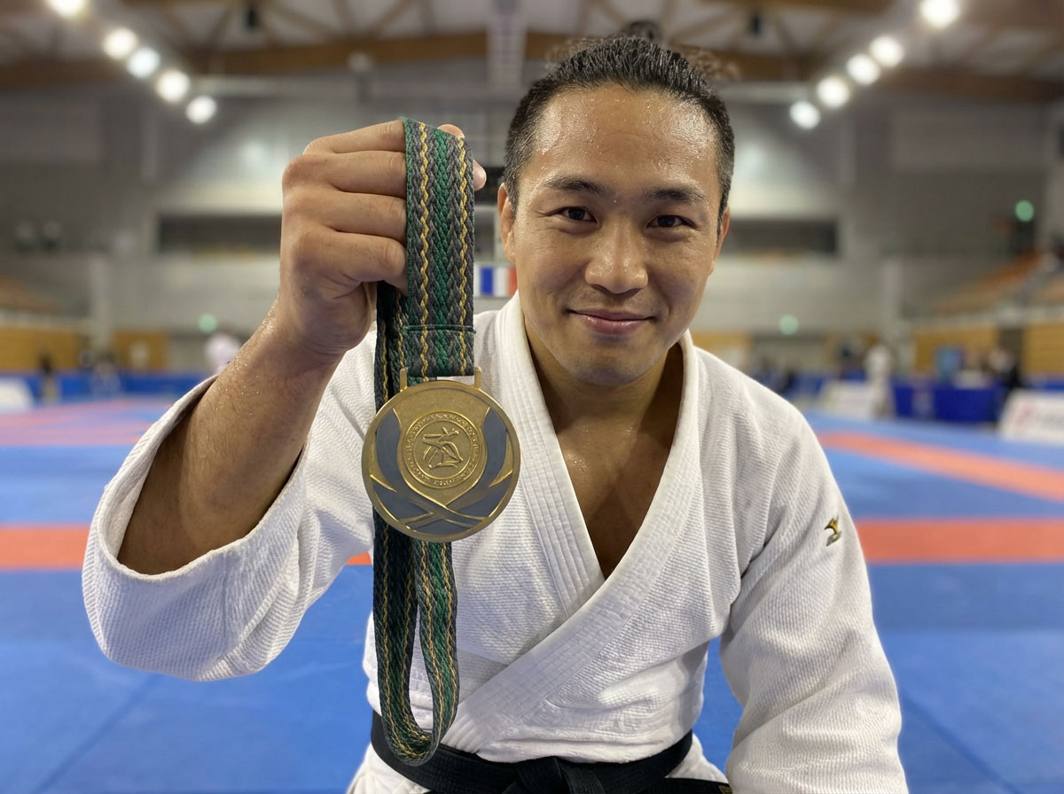 Martial artist in white gi smiling and proudly holding up a gold championship medal on the competition mat.