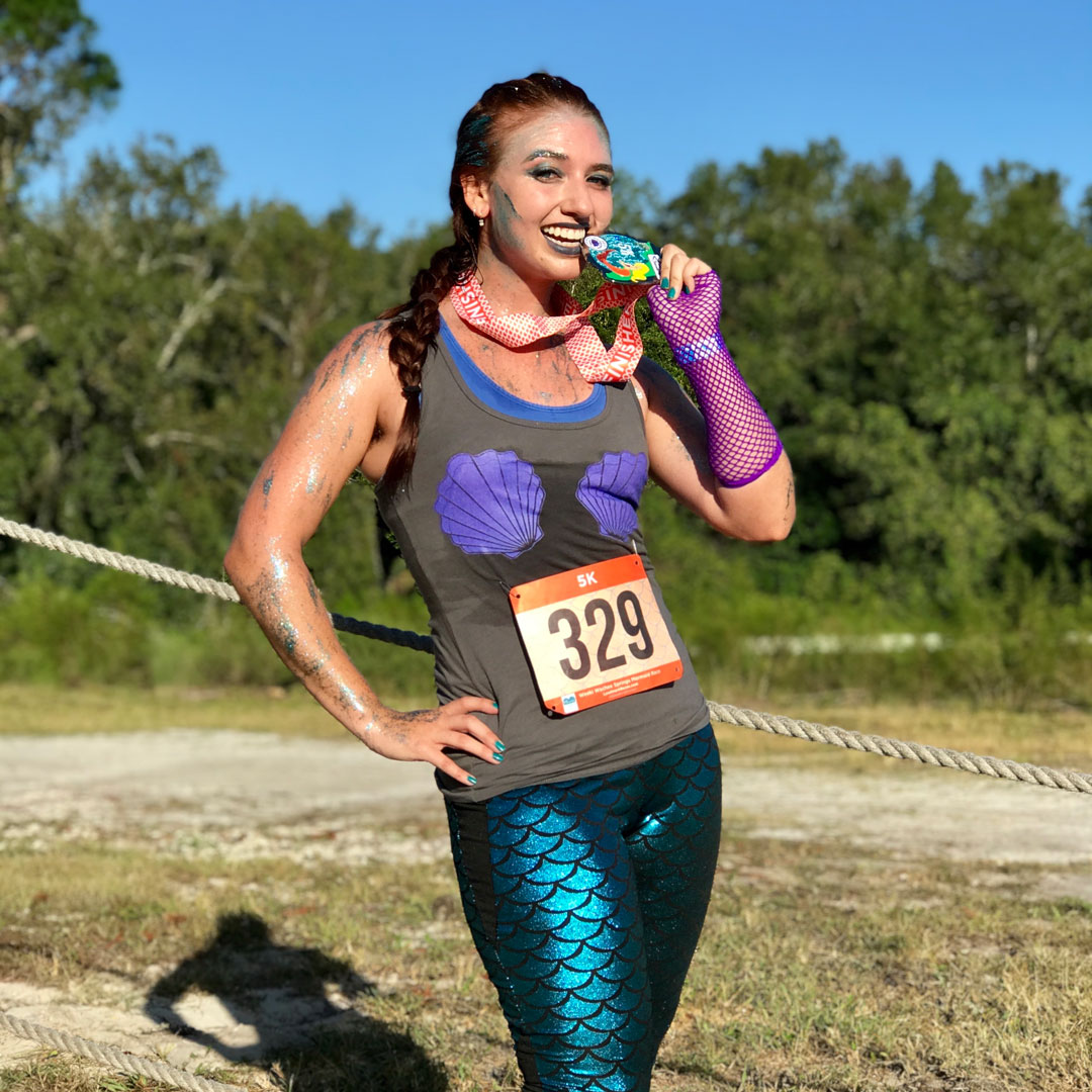 Runner sitting on a bench admiring her custom finisher medal with a red ribbon, with fellow runners chatting in the background.