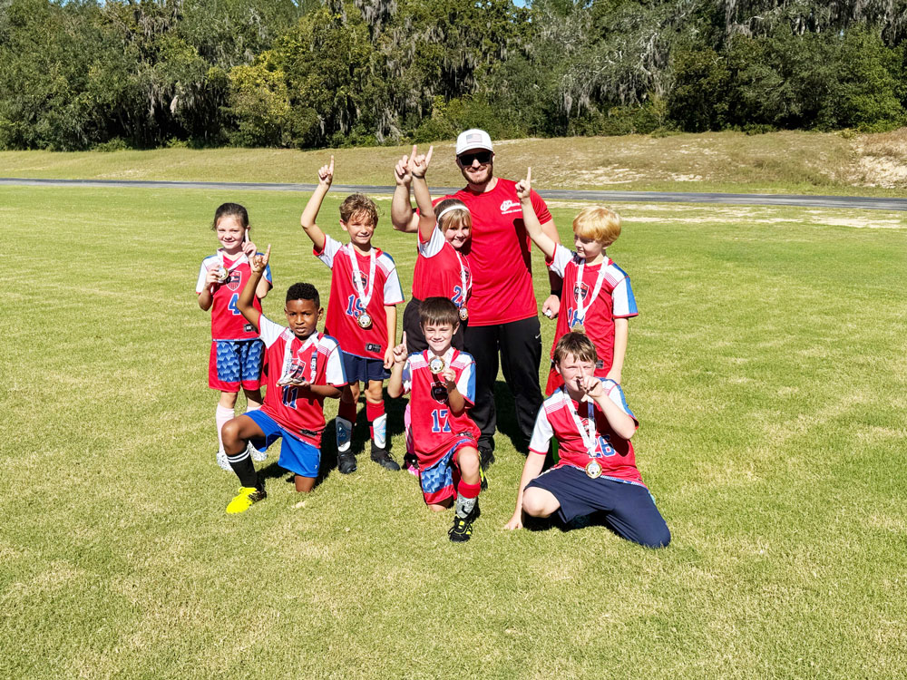 Youth soccer team posing with their coach on a grassy field, all wearing red and blue uniforms and proudly displaying medals, with some holding up one finger in celebration.