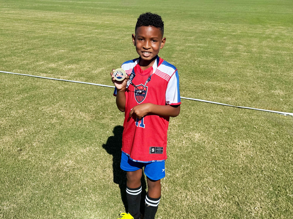 Young soccer player in a red and blue jersey smiling while holding up a medal on a sunny grassy field.