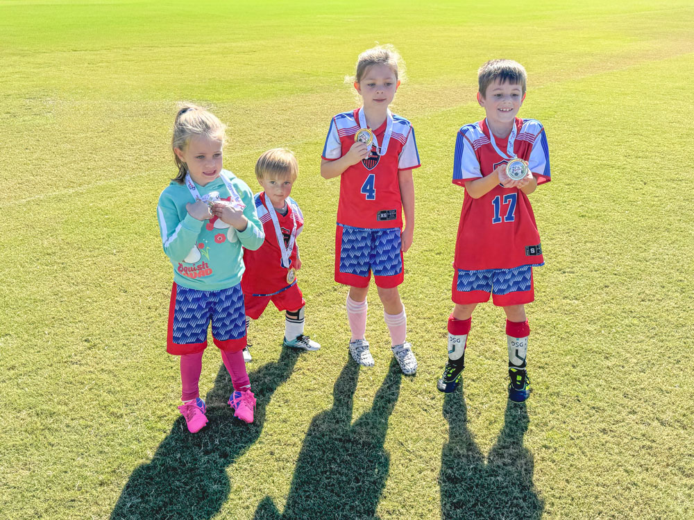 Four young children standing on a soccer field in bright sunlight, wearing red and blue soccer uniforms and proudly holding up medals, with one child in a mint green sweatshirt and pink socks.