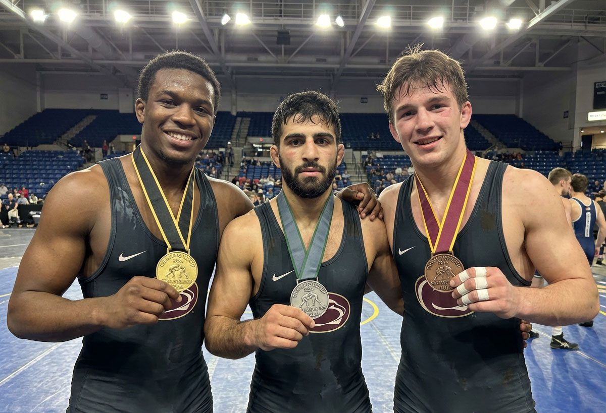 Three wrestlers in singlets holding up gold, silver, and bronze championship medals on the mat at a tournament arena.